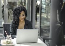 High angle of pensive African American female freelancer in glasses and casual clothes focusing on screen and interacting with netbook while sitting at table with glass of yummy drink on cafe terrace in sunny day